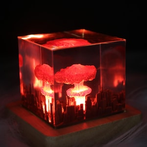 Peut inclure: Une lampe en résine rouge en forme de cube avec un nuage en forme de champignon et une ligne d'horizon de ville à l'intérieur. La lampe est éclairée de l'intérieur, projetant une lueur chaude.