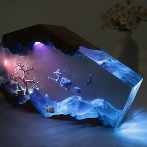 May include: A resin sculpture of a witch flying on a broomstick with a pumpkin and a tree in the background. The sculpture is illuminated from within with a blue and purple light.