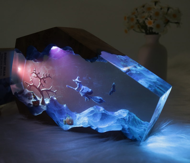 May include: A resin sculpture of a witch flying on a broomstick with a pumpkin and a tree in the background. The sculpture is illuminated from within with a blue and purple light.