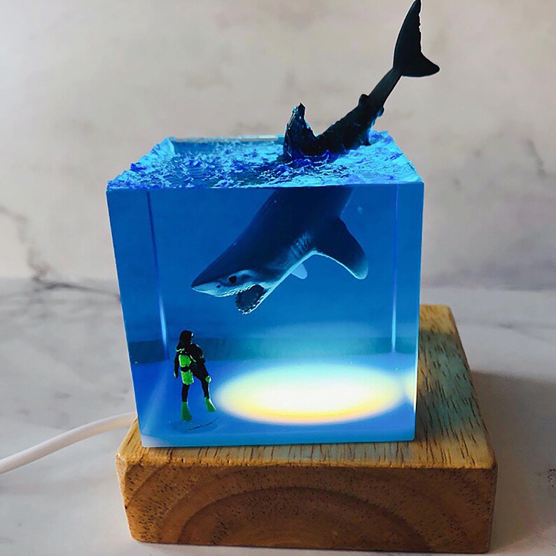 May include: A blue resin cube with a white shark and a diver figurine inside. The cube is lit from below with a warm white light and sits on a wooden base.