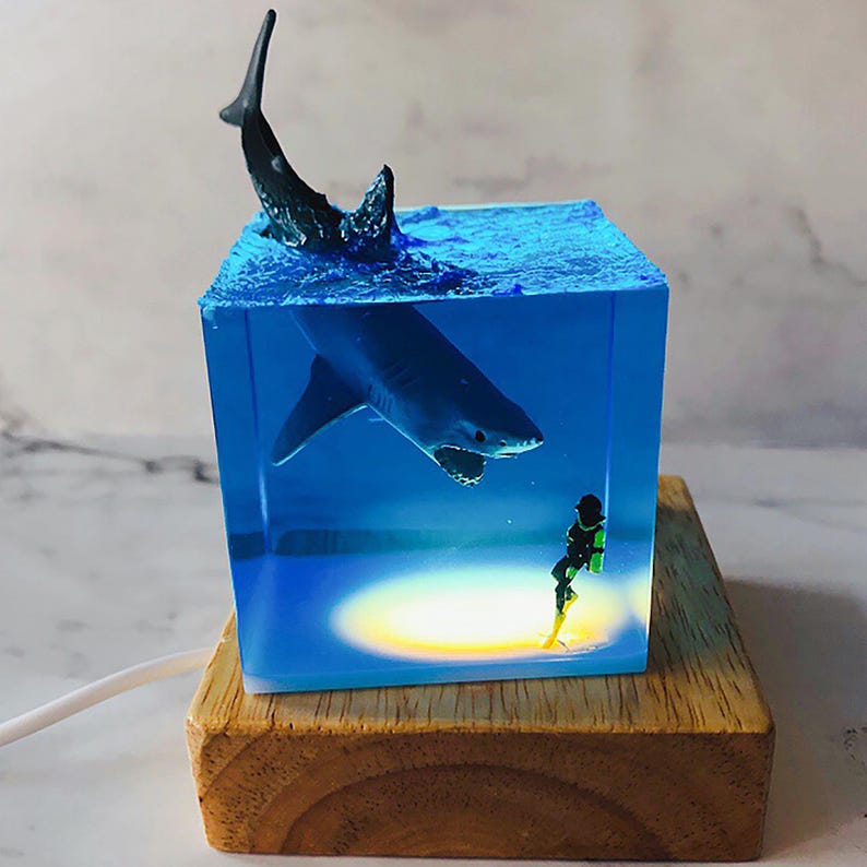 May include: A blue resin cube with a shark and a scuba diver inside. The cube is lit from below, casting a warm glow on the scene. The cube is sitting on a wooden base.