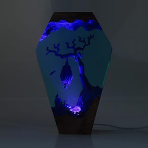 May include: A resin lamp shaped like a coffin with a blue glow. The lamp features a silhouette of a bat hanging from a tree branch inside the resin.