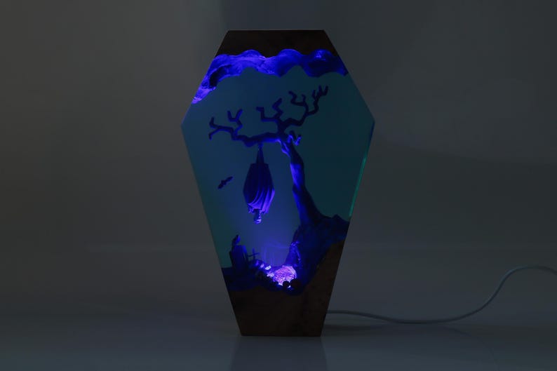 May include: A resin lamp shaped like a coffin with a blue glow. The lamp features a silhouette of a bat hanging from a tree branch inside the resin.