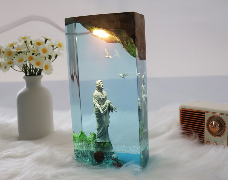 May include: A rectangular resin lamp with a wooden top and a clear resin body. The lamp features a figurine of a person standing in the water with birds flying above. The base of the lamp is filled with blue resin and has a green moss-like material at the bottom.