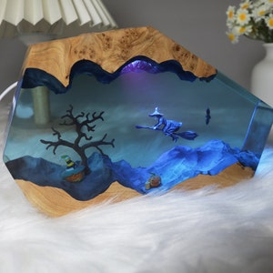 May include: A wooden resin lamp with a blue and purple resin scene inside. The scene includes a witch flying on a broomstick, a tree, and a pumpkin. The lamp is lit up with a blue light.