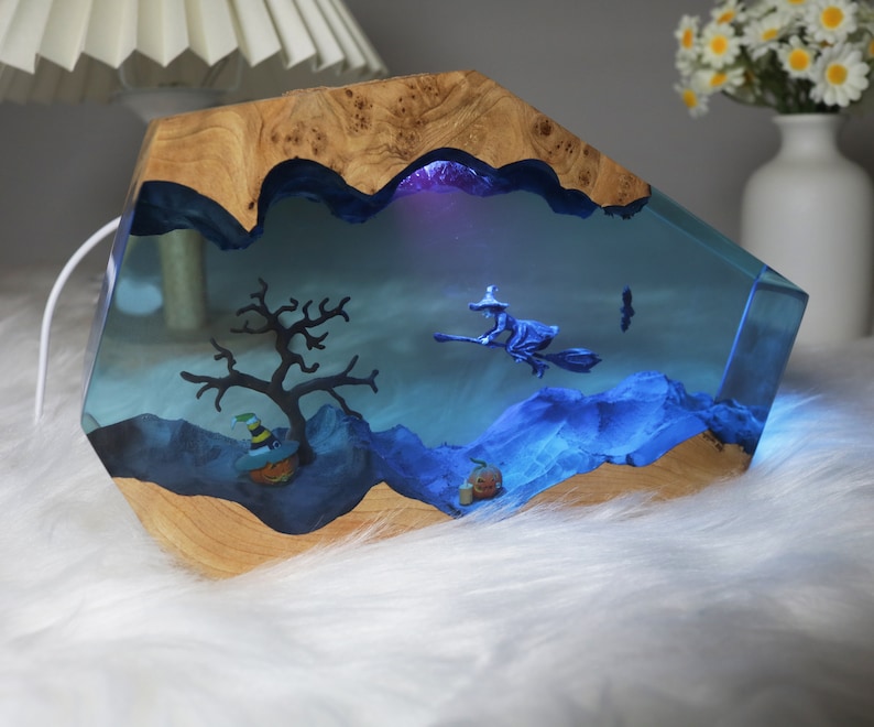 May include: A wooden resin lamp with a blue and purple resin scene inside. The scene includes a witch flying on a broomstick, a tree, and a pumpkin. The lamp is lit up with a blue light.