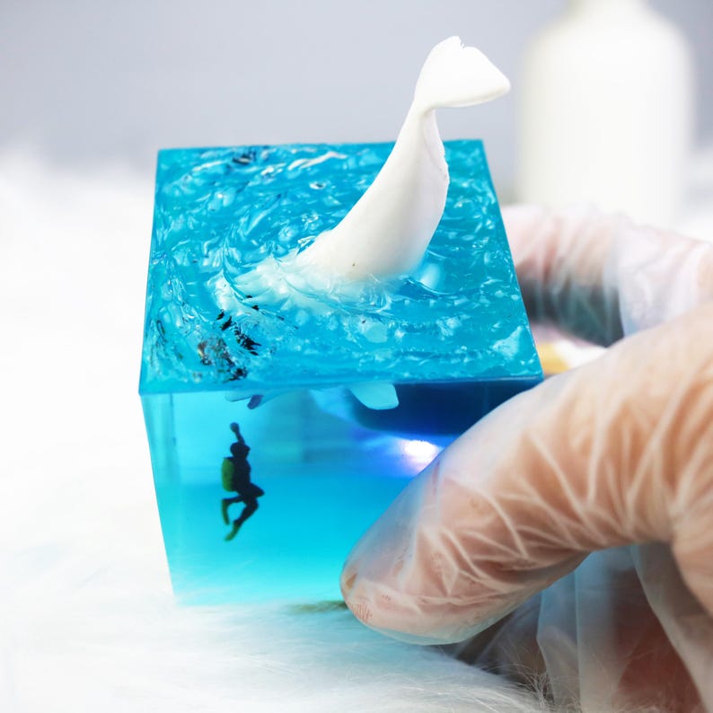 May include: A clear blue resin cube with a white whale figurine partially submerged in the water. A small black diver figurine is at the bottom of the cube.