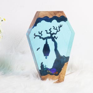 May include: A wooden coffin-shaped resin art piece with a blue and white background. A black silhouette of a tree with a bat hanging from a branch is depicted in the resin.