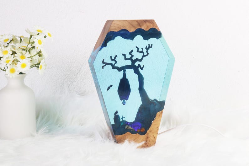 May include: A wooden coffin-shaped resin art piece with a blue and white background. A black silhouette of a tree with a bat hanging from a branch is depicted in the resin.