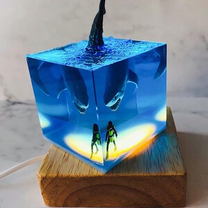 May include: A blue resin cube with a shark fin sticking out of the top. Inside the cube are two miniature scuba divers and a shark. The cube is lit from the bottom, casting a warm glow on the divers. The cube is sitting on a wooden base.
