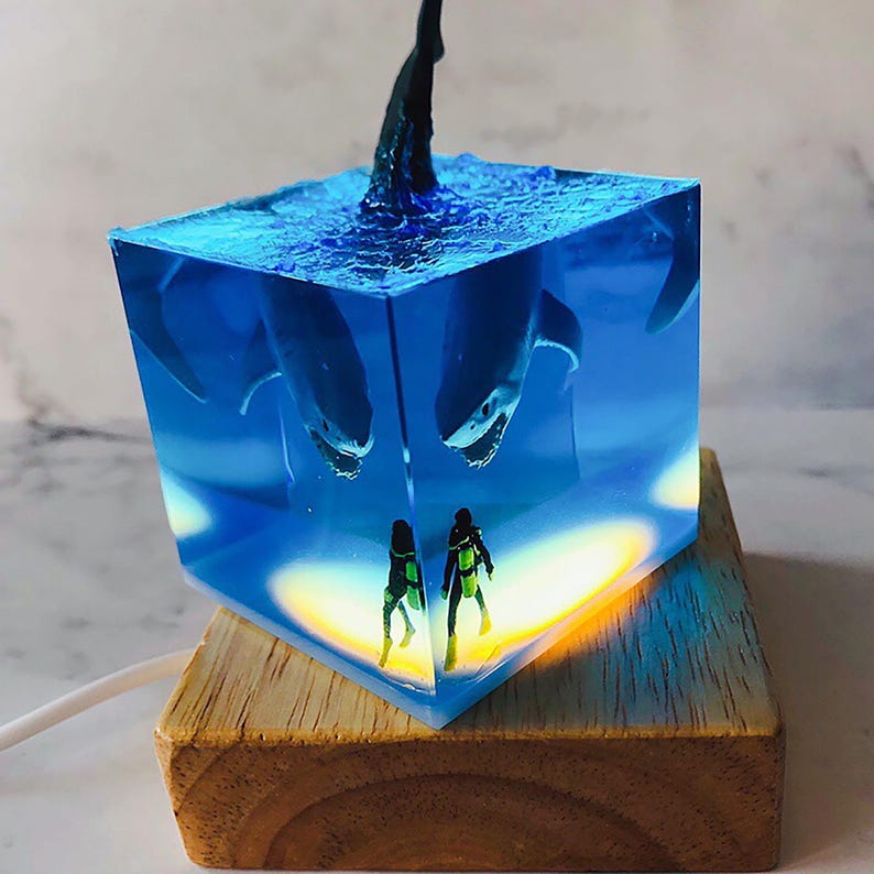 May include: A blue resin cube with a shark fin sticking out of the top. Inside the cube are two miniature scuba divers and a shark. The cube is lit from the bottom, casting a warm glow on the divers. The cube is sitting on a wooden base.