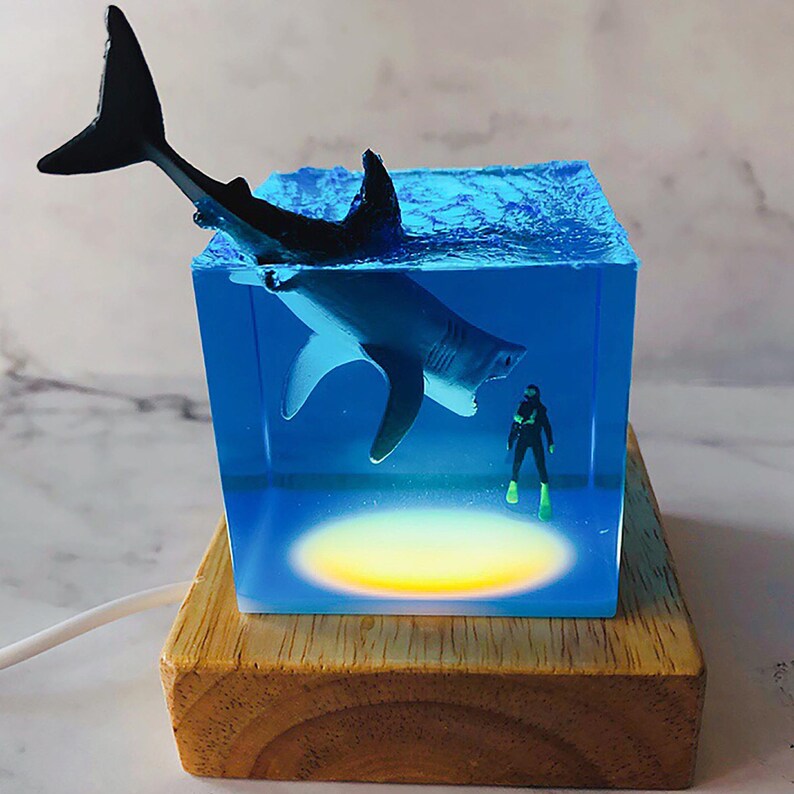 May include: A clear cube with a white shark figurine swimming inside. A miniature scuba diver figurine is at the bottom of the cube. The cube is lit from below with a warm yellow light and sits on a wooden base.