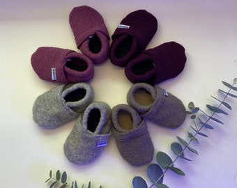 May include: A collection of baby booties in various shades of purple and gray, arranged in a circular pattern. The booties are made of a soft, felt-like material. Each pair has a small label with text. Eucalyptus leaves are in the background.