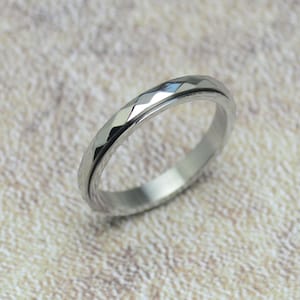 Könnte beinhalten: Ein silberfarbener Ring mit einem facettierten, geometrischen Design am äußeren Rand. Der Ring hat ein glattes, poliertes Innenband. Der Ring wird vor einem neutralen, strukturierten Hintergrund gezeigt.