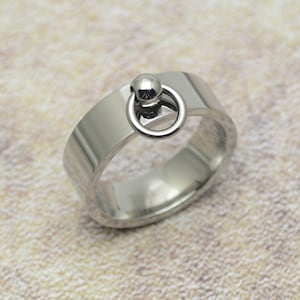 Könnte beinhalten: Ein silberner Ring mit einem runden, silbernen Ring, der oben angebracht ist. Der Ring ist dick und hat eine glatte, polierte Oberfläche.