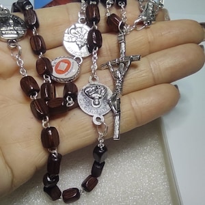 Op de afbeelding: Een bruine houten rozenkrans met zilveren kralen en een zilveren kruisbeeld. De rozenkrans zit in een witte doos met een afbeelding van paus Johannes Paulus II op de deksel.