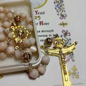 May include: A rosary with pale pink beads and gold-toned metal accents, including a crucifix and a centrepiece featuring a figure. The rosary is partially contained in a white plastic case next to a card with the text Year 25 of Hope.