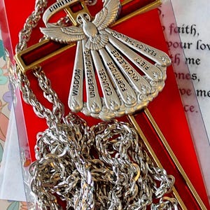 Peut inclure: Un collier de confirmation argenté avec un pendentif en forme de croix et une chaîne. Le pendentif représente une colombe et les mots "Sagesse", "Compréhension", "Conseil", "Force", "Connaissance", "Piété" et "Crainte du Seigneur". La croix est rouge avec des bordures dorées.