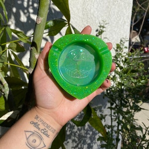 Puede incluir: Un cenicero verde con purpurina en forma de hoja con un interior transparente y translúcido. El cenicero tiene un diseño de hongo con un contorno blanco.