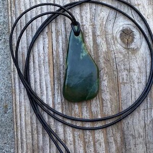 May include: A dark green jade pendant on a black leather cord. The pendant is a freeform shape and has a smooth, polished surface.
