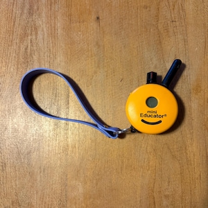 May include: An orange Mini Educator® dog training collar with a black antenna and a purple strap. The collar has a small screen and the brand name printed on the front. The collar is designed for dog training.