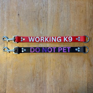 以下が含まれることがあります： 2つの犬用リードアタッチメント。1つは赤色で、白文字で「WORKING K9」と書かれています。もう1つは黒色で、紫色の文字で「DO NOT PET」と書かれています。どちらもシルバーの金属製留め金とDリングが付いています。