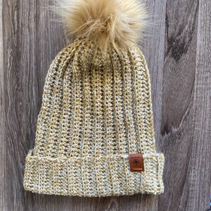 May include: A beige and white crocheted beanie with a fluffy brown pom pom on top. The beanie has a brown leather patch with the word "Nomad" and a compass symbol.
