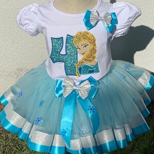 May include: A white t-shirt with a blue and glitter number "4" and a glittery image of Elsa from Frozen. The shirt is paired with a light blue tulle tutu with silver and blue ribbon accents and snowflake designs.