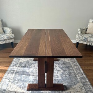 May include: A dark brown wooden dining table with a split top and a unique base design. The table is set on a gray rug in a room with hardwood floors.