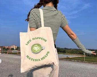 May include: Beige tote bag with the text "LIFE HAPPENS MATCHA HELPS" and a green matcha latte design. The bag is carried over the shoulder. The person is wearing a green and white striped shirt and light-colored pants.