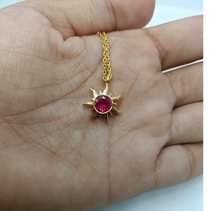 May include: A gold necklace with a sun-shaped pendant, featuring a central red gemstone. The pendant is attached to a fine chain. The necklace is shown against a light background.