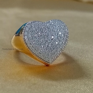 Pode incluir: Um anel dourado com uma face em forma de coração coberta por pequenas pedras brilhantes. O anel está sobre um fundo dourado suave. O nome da marca "Trinity Gold & Diamonds" é visível.