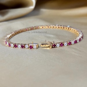 Op de afbeelding: Een gouden tennisarmband met afwisselende rode en witte ronde stenen. De armband is open en de sluiting is zichtbaar.