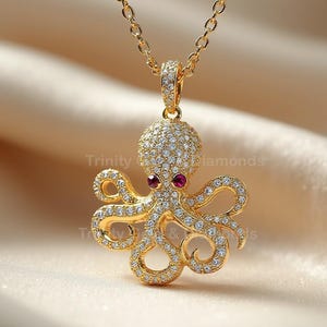 May include: A gold-colored octopus pendant necklace. The octopus is encrusted with small, clear gemstones and has two red gemstone eyes. The pendant hangs from a delicate gold chain. The background is a soft, neutral fabric.