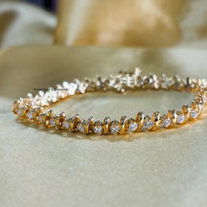 Puede incluir: Un brazalete de oro con una fila de piedras redondas transparentes engastadas en zigzag. El brazalete est&aacute; sobre un fondo de tela dorada.