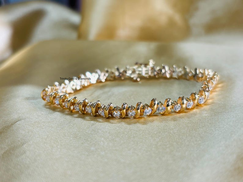 Puede incluir: Un brazalete de oro con una fila de piedras redondas transparentes engastadas en zigzag. El brazalete est&aacute; sobre un fondo de tela dorada.