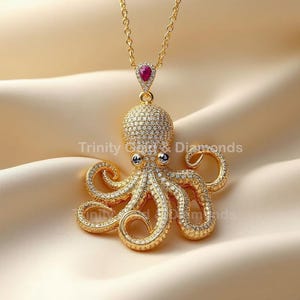 Op de afbeelding: Een gouden ketting met een hanger in de vorm van een octopus met een sprankelend, met edelstenen bezet ontwerp. De octopus heeft meerdere tentakels en een ronde kop. Een druppelvormige rode edelsteen hangt boven de octopus. De tekst "Trinity Gold & Diamonds" is zichtbaar.