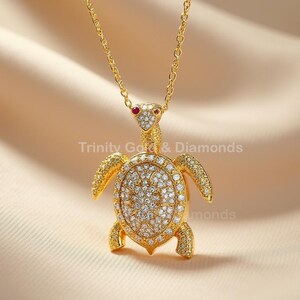 Op de afbeelding: Een goudkleurige schildpadhangerketting. De schildpadhanger is bezet met kleine, sprankelende stenen en heeft rode accenten voor de ogen. De ketting wordt gepresenteerd op een beige stoffen achtergrond. De tekst "Trinity Gold & Diamonds" is zichtbaar.