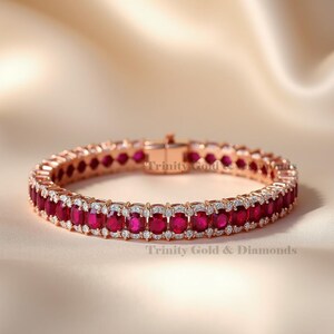 Puede incluir: Pulsera de oro rosa con piedras preciosas de rubí de corte ovalado y pequeños detalles de diamantes. La pulsera tiene un cierre seguro y está inscrita con "Trinity Gold & Diamonds". La joya se exhibe sobre una superficie satinada.