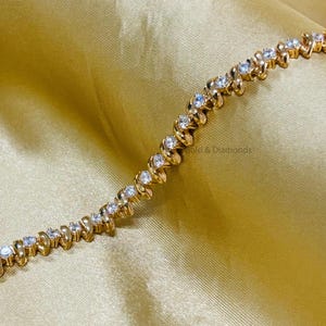 Puede incluir: Un brazalete de oro con un dise&ntilde;o ondulado y piedras redondas transparentes. El brazalete est&aacute; sobre un fondo de tela dorada.