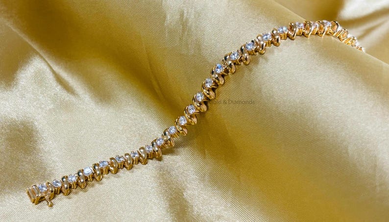 Puede incluir: Un brazalete de oro con un dise&ntilde;o ondulado y piedras redondas transparentes. El brazalete est&aacute; sobre un fondo de tela dorada.