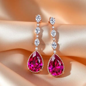 Könnte beinhalten: Ohrringe zum Hängen in Roségold. Jeder Ohrring hat einen großen, birnenförmigen, leuchtend pinkfarbenen Edelstein am unteren Ende, der von kleineren weißen Edelsteinen und Steinen im Marquiseschliff akzentuiert wird. Die Ohrringe werden auf einem Satinhintergrund präsentiert.