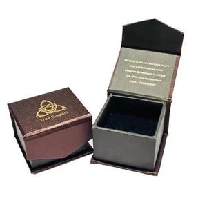 Puede incluir: Una caja de anillo de terciopelo marr&oacute;n con un logotipo dorado que dice "True Elegant" y un dise&ntilde;o de nudo celta. La caja est&aacute; abierta y el interior est&aacute; forrado de terciopelo negro.