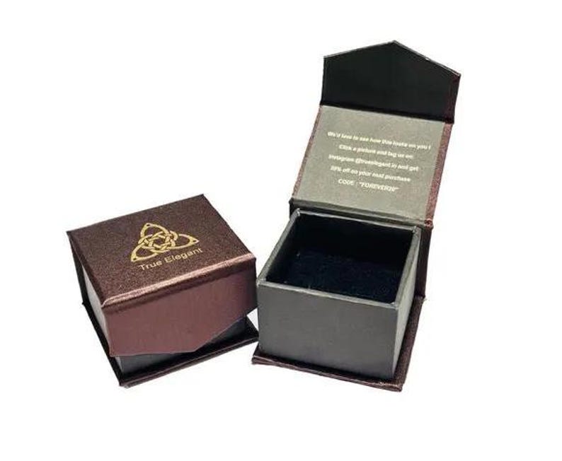 Puede incluir: Una caja de anillo de terciopelo marr&oacute;n con un logotipo dorado que dice "True Elegant" y un dise&ntilde;o de nudo celta. La caja est&aacute; abierta y el interior est&aacute; forrado de terciopelo negro.