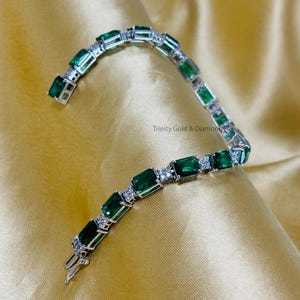 May include: A silver bracelet with emerald green and clear rectangular stones. The bracelet is on a gold fabric background.