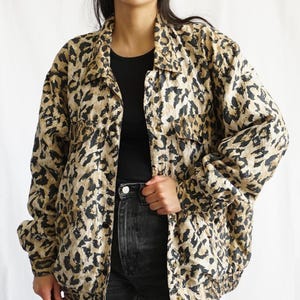 Könnte beinhalten: Eine Jacke mit Leopardenmuster in Beige und Schwarz, mit Kragen, zwei Brusttaschen und Knopfverschluss. Die Jacke hat lange Ärmel und einen gerippten Saum. Das Model trägt ein schwarzes Oberteil und eine schwarze Jeans.