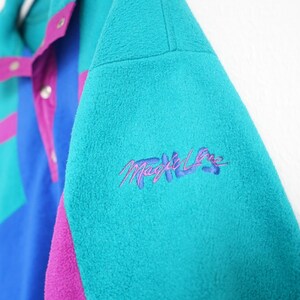 Op de afbeelding: Een turquoise, blauw en paars fleecejack met de geborduurde tekst "Magic Lane" op de mouw. De jas heeft een colorblock-ontwerp met drukknoopsluitingen en een hoge kraag. Het fleecemateriaal ziet er zacht en warm uit.