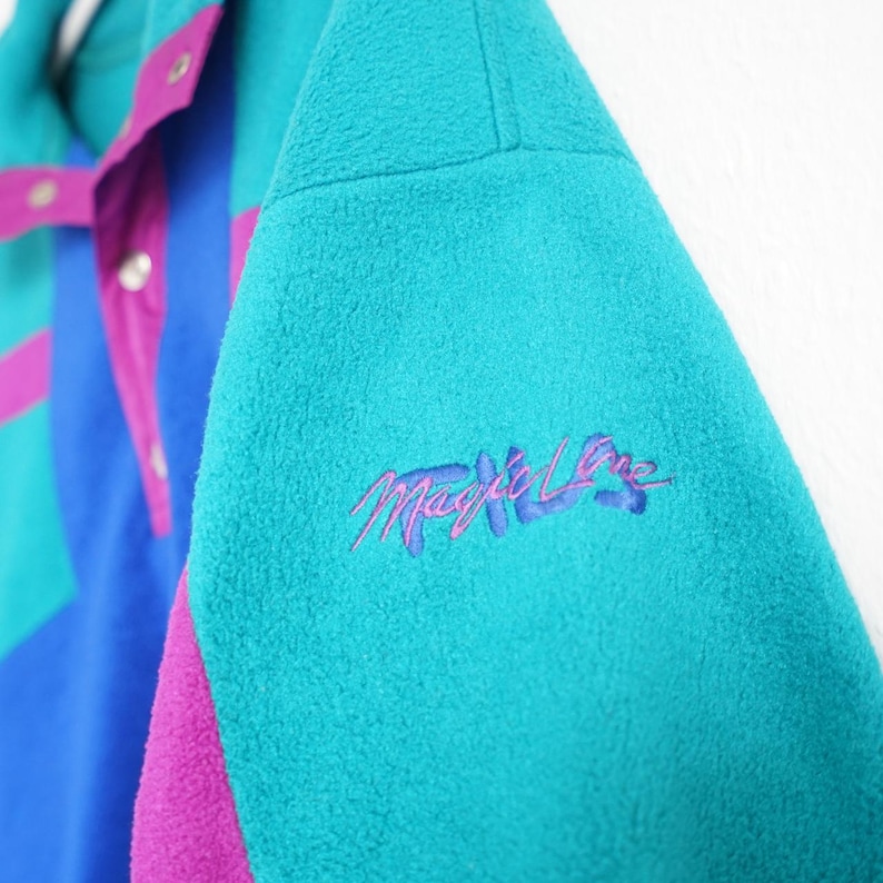 Op de afbeelding: Een turquoise, blauw en paars fleecejack met de geborduurde tekst "Magic Lane" op de mouw. De jas heeft een colorblock-ontwerp met drukknoopsluitingen en een hoge kraag. Het fleecemateriaal ziet er zacht en warm uit.
