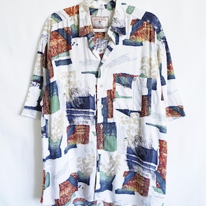 Könnte beinhalten: Weißes Button-Down-Hemd mit einem farbenfrohen abstrakten Muster. Das Hemd hat kurze Ärmel und einen Kragen. Das Design umfasst rechteckige Formen in Rot-, Blau-, Grün- und Beigetönen. Das Hemd hängt an einem weißen Kleiderbügel.