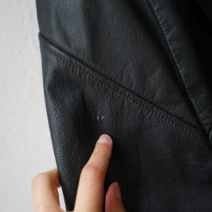 May include: Close-up of a black leather jacket. The jacket has visible stitching and a textured surface. A finger is touching the jacket, highlighting the material's details. The jacket appears to be well-worn.